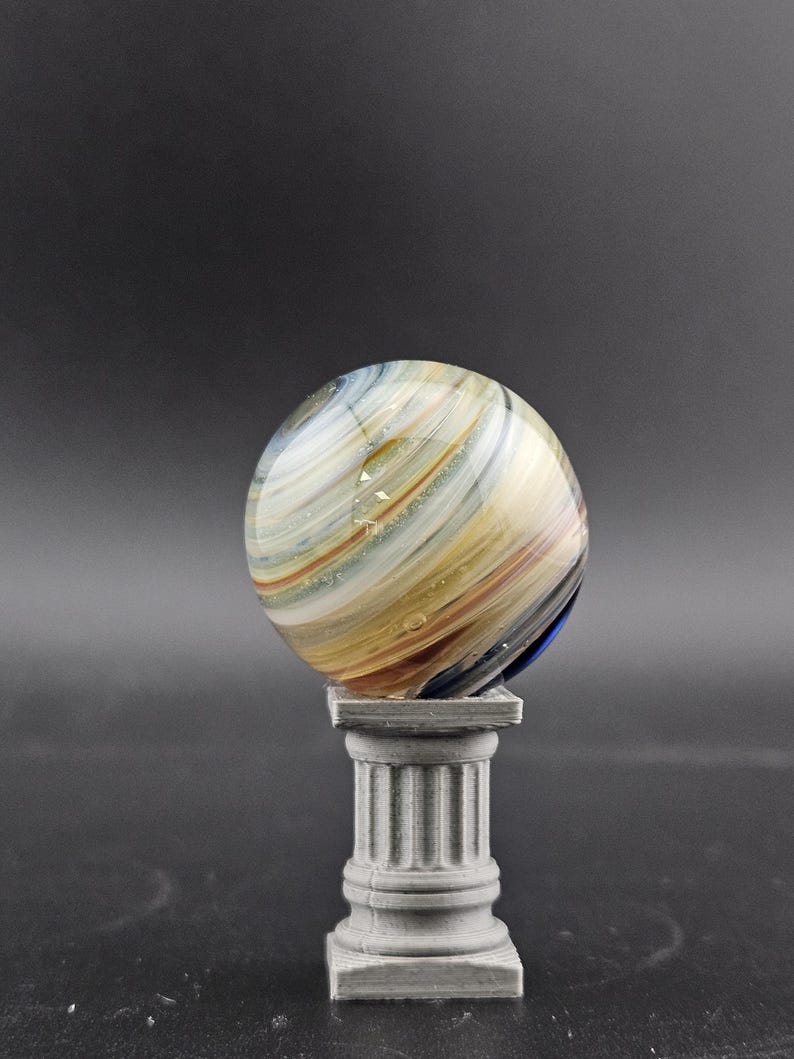 May include: A colorful glass marble with swirling patterns of white, yellow, and blue rests on a small, gray, classical-style pedestal. The marble's surface is reflective, and the pedestal has a fluted column design.