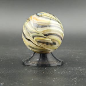 May include: A decorative glass marble with swirling patterns of yellow, black, and white, resting on a black, ridged base. The marble is spherical and appears to be handmade, with a unique design. The base is circular and provides a stable foundation.