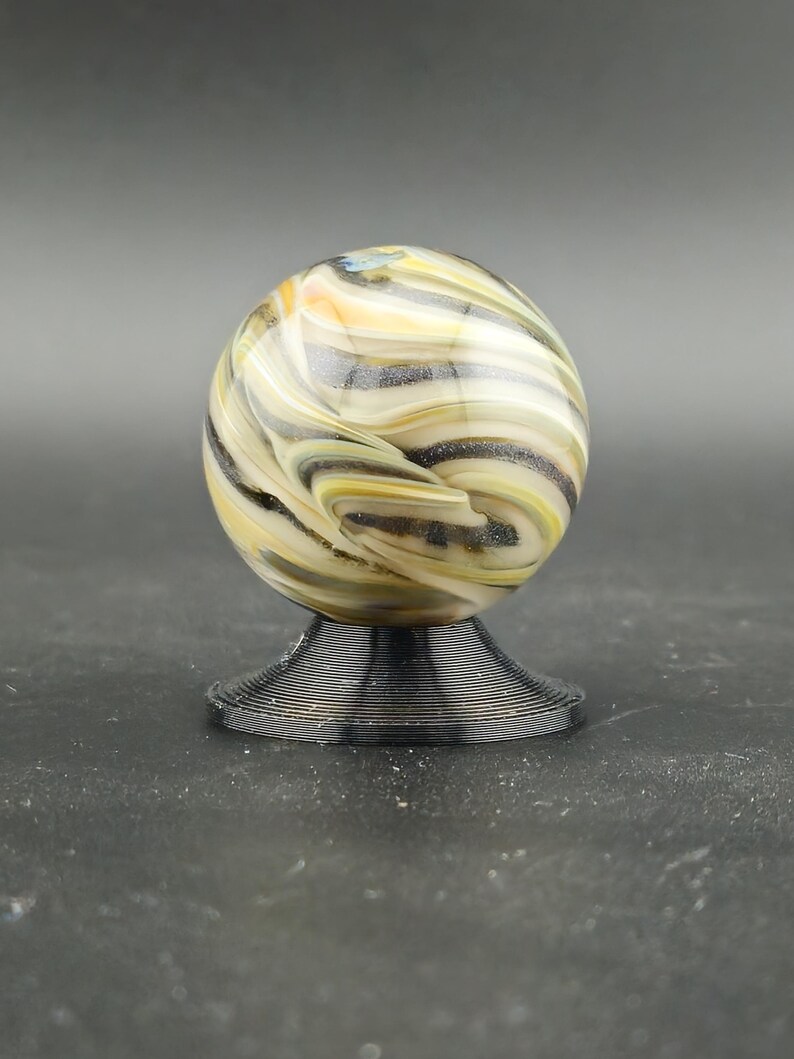 May include: A decorative glass marble with swirling patterns of yellow, black, and white, resting on a black, ridged base. The marble is spherical and appears to be handmade, with a unique design. The base is circular and provides a stable foundation.