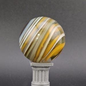 May include: A decorative glass marble with swirling patterns of yellow, white, and brown, resting on a small, gray, classical-style pedestal. The marble is spherical and appears to be made of glass.