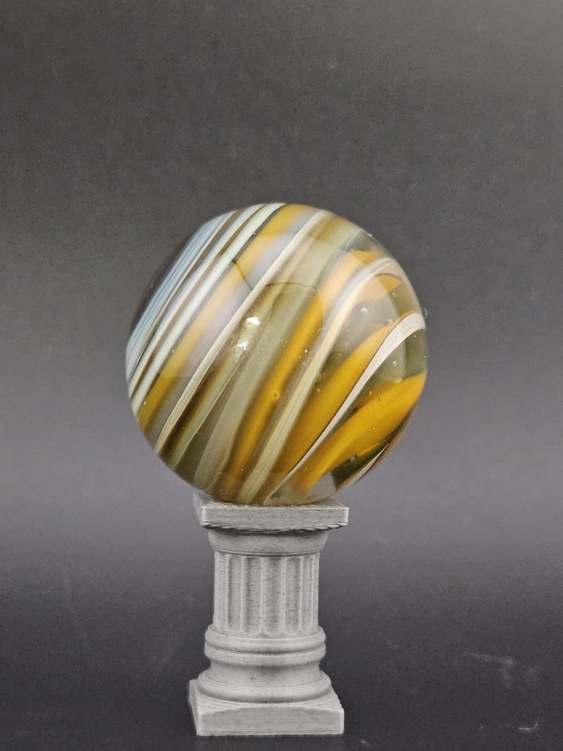 May include: A decorative glass marble with swirling patterns of yellow, white, and brown, resting on a small, gray, classical-style pedestal. The marble is spherical and appears to be made of glass.