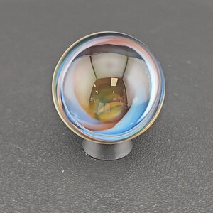 May include: A spherical glass marble featuring swirling internal patterns of blue, white, and amber hues. The clear glass surface reflects light, creating a glossy appearance. The marble is supported by a small, circular, dark gray plastic stand.