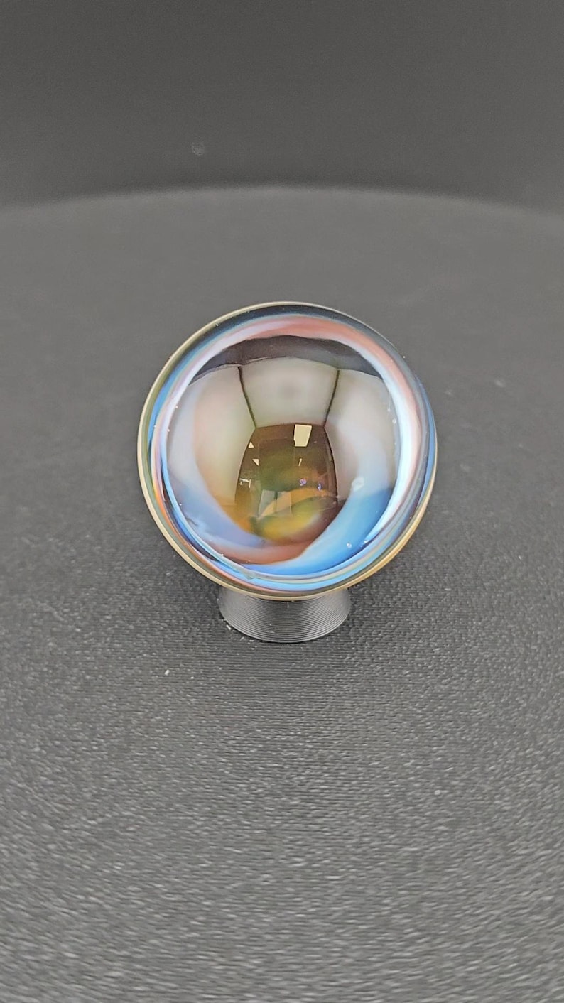 May include: A spherical glass marble featuring swirling internal patterns of blue, white, and amber hues. The clear glass surface reflects light, creating a glossy appearance. The marble is supported by a small, circular, dark gray plastic stand.