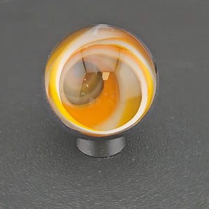 May include: A spherical glass marble featuring a clear outer layer with internal swirling patterns of opaque white and vibrant orange colors. The marble is resting on a small, dark, circular stand, showcasing a smooth, polished surface.