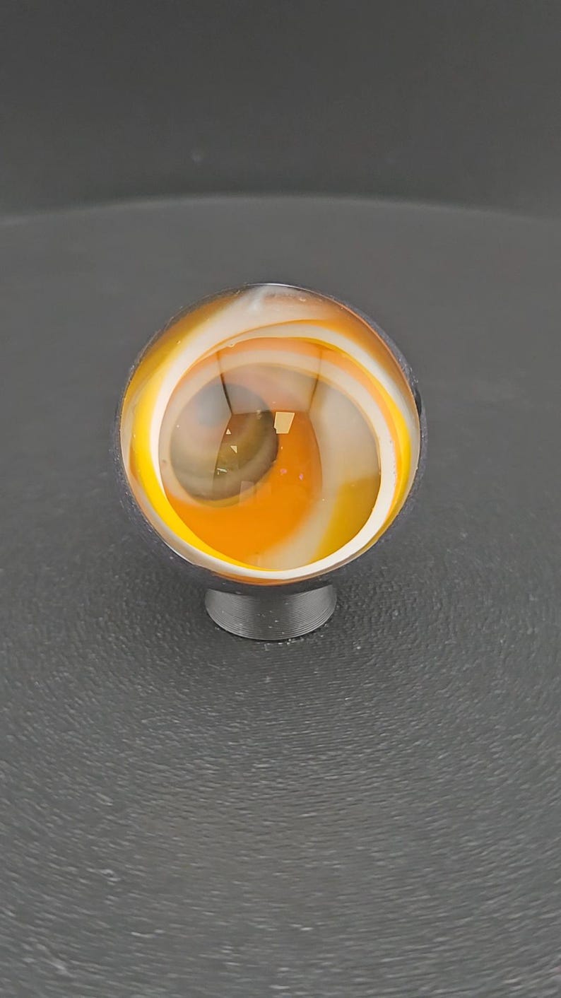 May include: A spherical glass marble featuring a clear outer layer with internal swirling patterns of opaque white and vibrant orange colors. The marble is resting on a small, dark, circular stand, showcasing a smooth, polished surface.