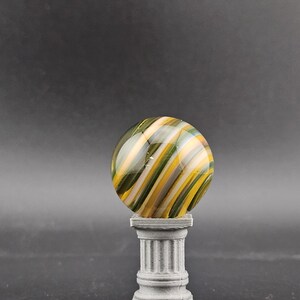 May include: A decorative glass marble with green, yellow, and white swirling stripes, displayed on a small, gray, classical-style pedestal. The marble is spherical and the pedestal has a fluted column design.