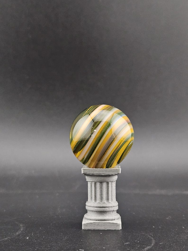 May include: A decorative glass marble with green, yellow, and white swirling stripes, displayed on a small, gray, classical-style pedestal. The marble is spherical and the pedestal has a fluted column design.