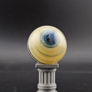 May include: A decorative glass sphere with a swirling pattern of yellow, blue, and black, resting on a gray, miniature column. The sphere has a polished, reflective surface, and the column has a classical design.