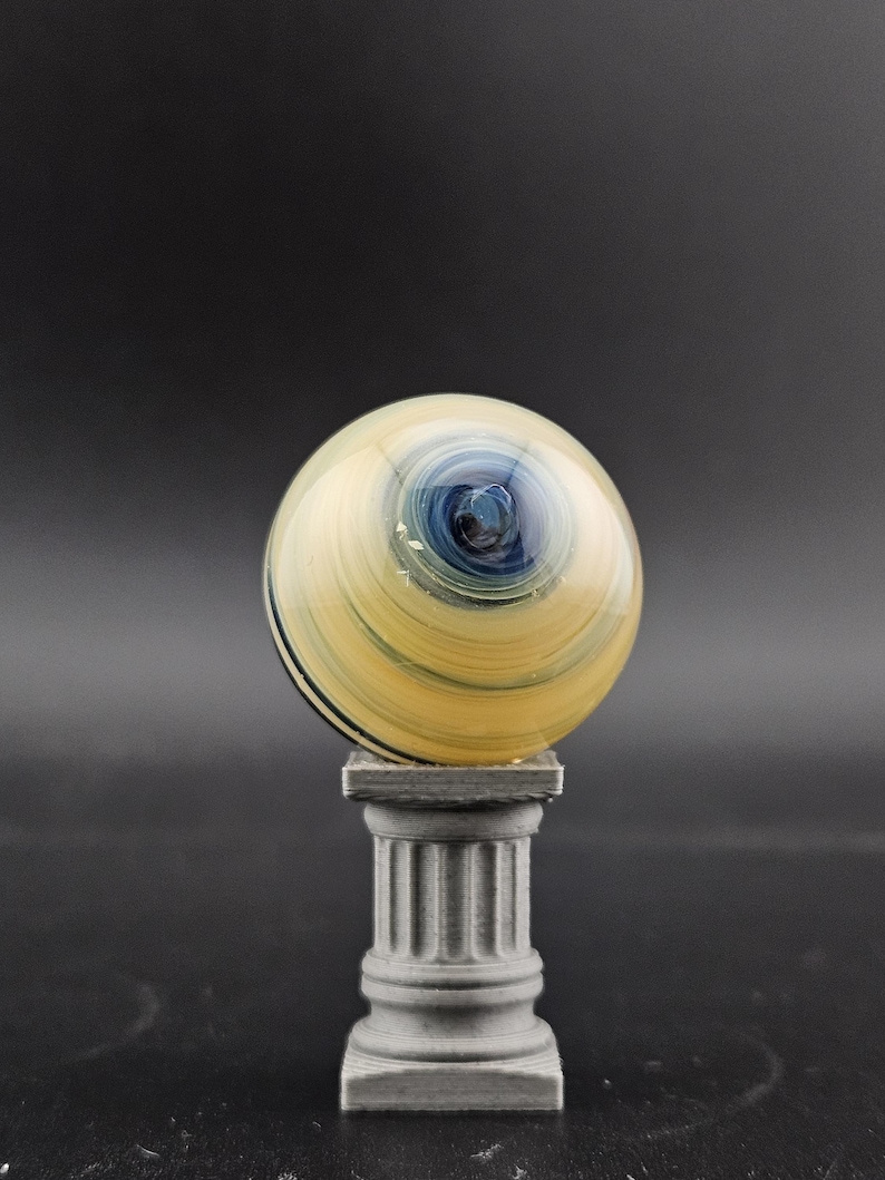 May include: A decorative glass sphere with a swirling pattern of yellow, blue, and black, resting on a gray, miniature column. The sphere has a polished, reflective surface, and the column has a classical design.