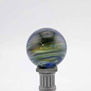 May include: A decorative glass sphere with swirling blue, green, and yellow colors, resting on a small, gray, classical-style pedestal. The sphere has a glossy finish and appears to be made of glass.
