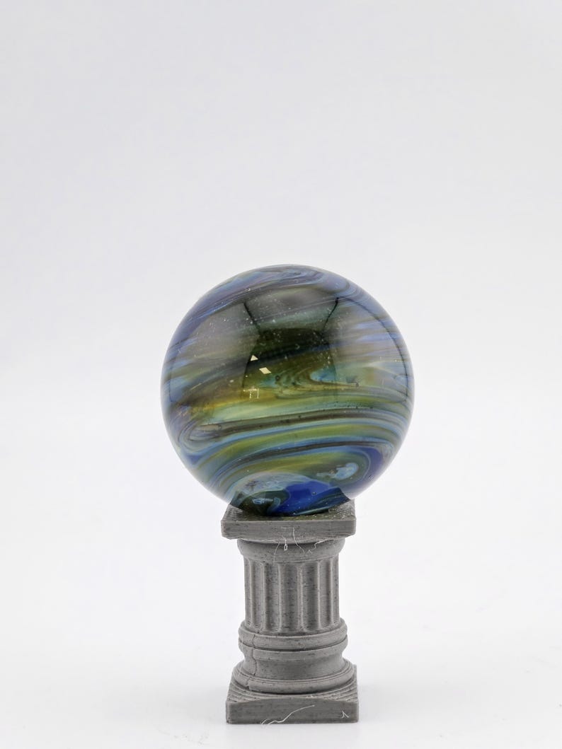 May include: A decorative glass sphere with swirling blue, green, and yellow colors, resting on a small, gray, classical-style pedestal. The sphere has a glossy finish and appears to be made of glass.