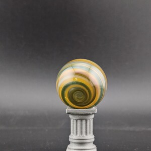 May include: A decorative glass sphere with a swirling pattern of green, yellow, and orange hues. The sphere rests atop a small, gray, classical-style pedestal. The background is a solid, dark gray.