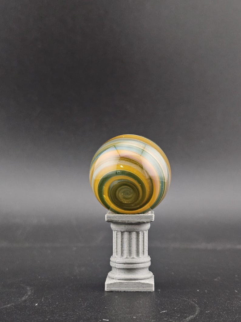 May include: A decorative glass sphere with a swirling pattern of green, yellow, and orange hues. The sphere rests atop a small, gray, classical-style pedestal. The background is a solid, dark gray.