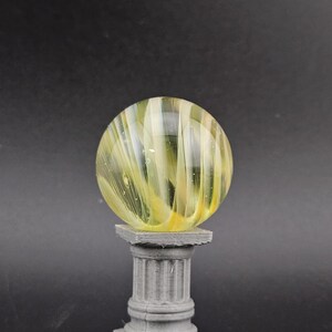 May include: A decorative glass marble with yellow and white swirls, resting on a gray, fluted column pedestal. The marble is spherical and appears translucent, with light passing through the glass. The pedestal has a square base and top.