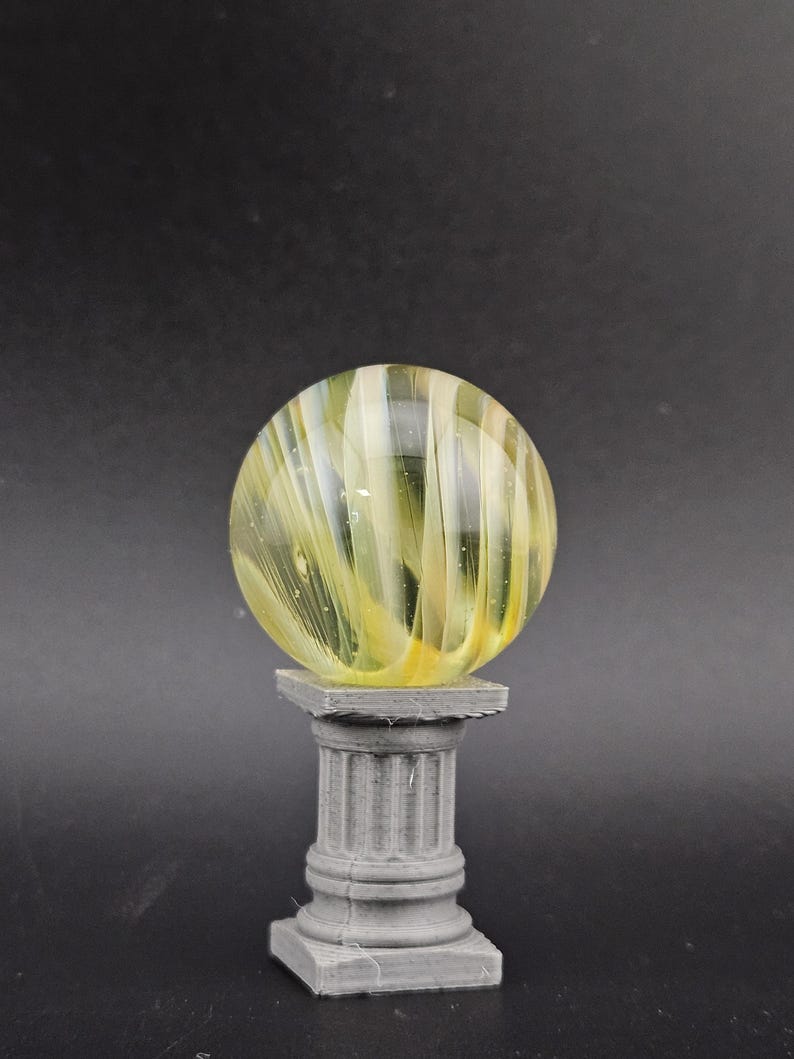 May include: A decorative glass marble with yellow and white swirls, resting on a gray, fluted column pedestal. The marble is spherical and appears translucent, with light passing through the glass. The pedestal has a square base and top.