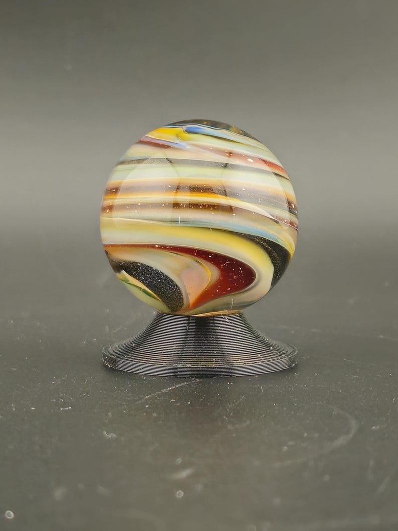 May include: A spherical glass marble featuring swirling bands of yellow, white, brown, and black colors with subtle glittery flecks throughout. The marble is resting on a small, circular, black, ribbed plastic display stand.