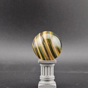 May include: A decorative glass marble with yellow, green, and white stripes, resting on a small, gray, classical-style pedestal. The marble is spherical and appears to be made of glass. The pedestal has a fluted column design.
