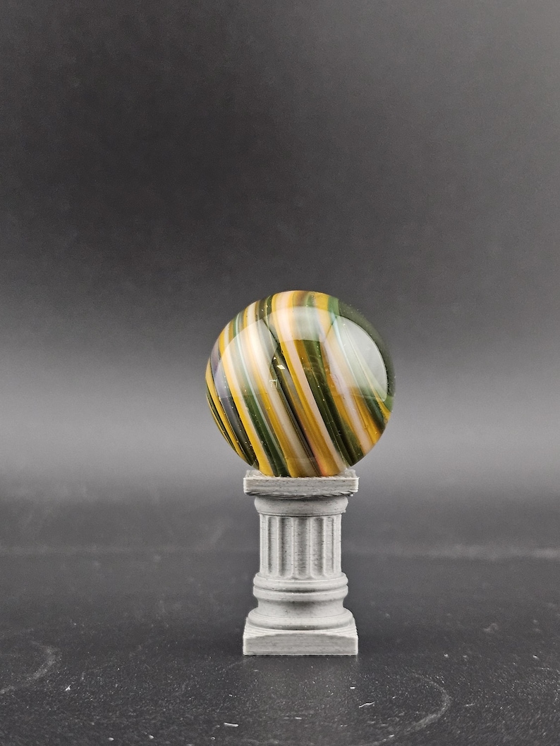 May include: A decorative glass marble with yellow, green, and white stripes, resting on a small, gray, classical-style pedestal. The marble is spherical and appears to be made of glass. The pedestal has a fluted column design.