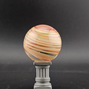 May include: A decorative glass sphere with swirling orange, yellow, and tan colors, resting on a gray, classical-style pedestal. The sphere appears to be made of layered glass, creating a unique striped pattern. The pedestal has a fluted column design.
