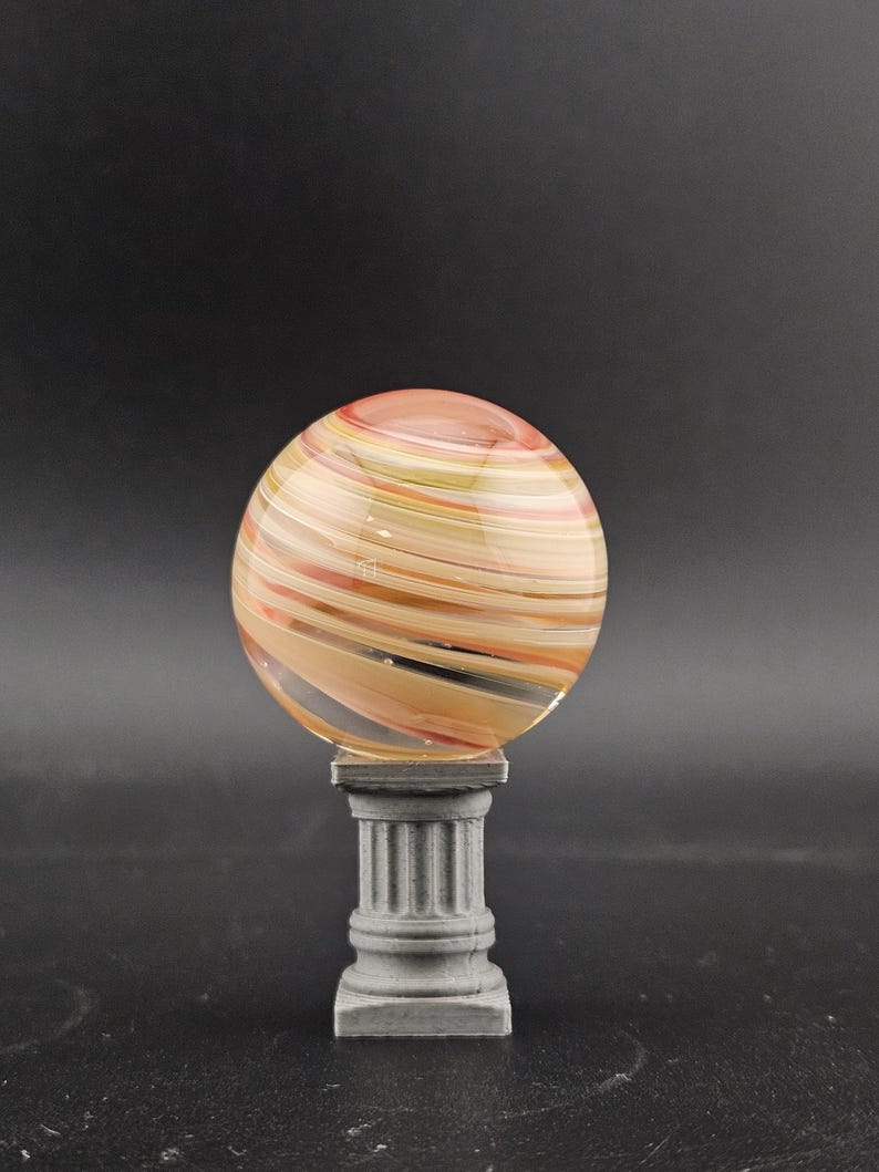 May include: A decorative glass sphere with swirling orange, yellow, and tan colors, resting on a gray, classical-style pedestal. The sphere appears to be made of layered glass, creating a unique striped pattern. The pedestal has a fluted column design.