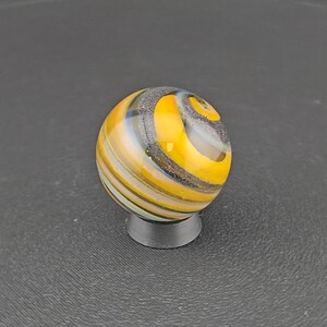 May include: A spherical glass marble featuring swirling patterns of bright yellow, translucent white, and dark grey with a subtle glittery texture. The marble is resting on a small, circular, dark grey stand that supports its rounded base.