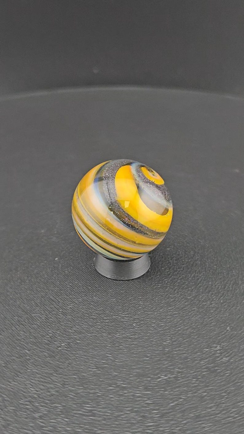 May include: A spherical glass marble featuring swirling patterns of bright yellow, translucent white, and dark grey with a subtle glittery texture. The marble is resting on a small, circular, dark grey stand that supports its rounded base.
