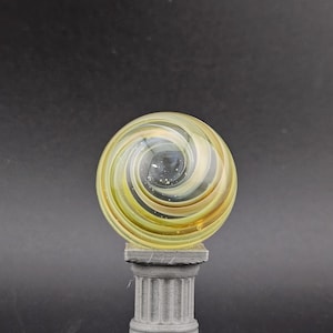 May include: A decorative glass marble with a yellow and brown spiral design, resting on a gray, fluted column. The marble has a swirling pattern and is set against a dark background.
