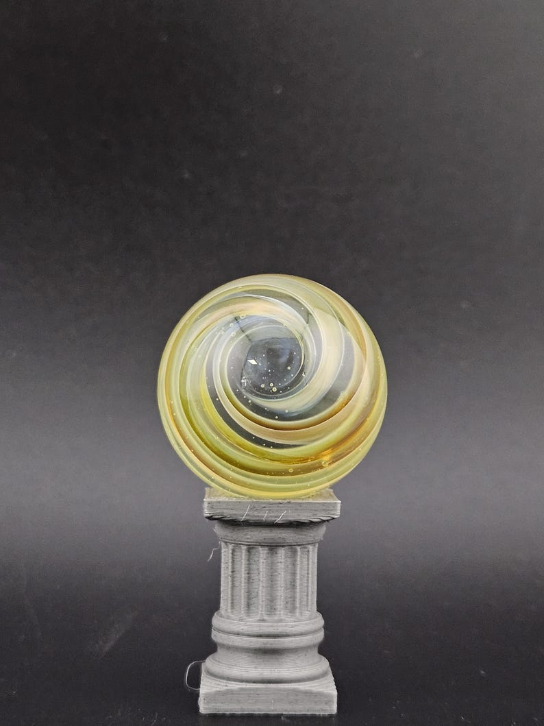 May include: A decorative glass marble with a yellow and brown spiral design, resting on a gray, fluted column. The marble has a swirling pattern and is set against a dark background.