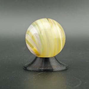 May include: A yellow and white swirled glass marble sits atop a black, spiral-shaped stand. The marble has a glossy finish and is illuminated, showcasing the intricate patterns within the glass. The stand provides a stable base for the decorative sphere.