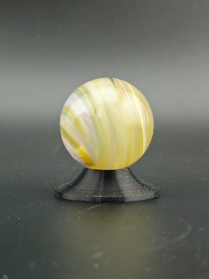 May include: A yellow and white swirled glass marble sits atop a black, spiral-shaped stand. The marble has a glossy finish and is illuminated, showcasing the intricate patterns within the glass. The stand provides a stable base for the decorative sphere.