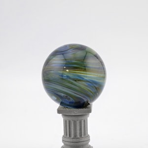 May include: A decorative glass orb with swirling blue, green, and yellow colors, resting on a small, gray, classical-style column. The orb has a polished, reflective surface, and the column has fluted detailing.