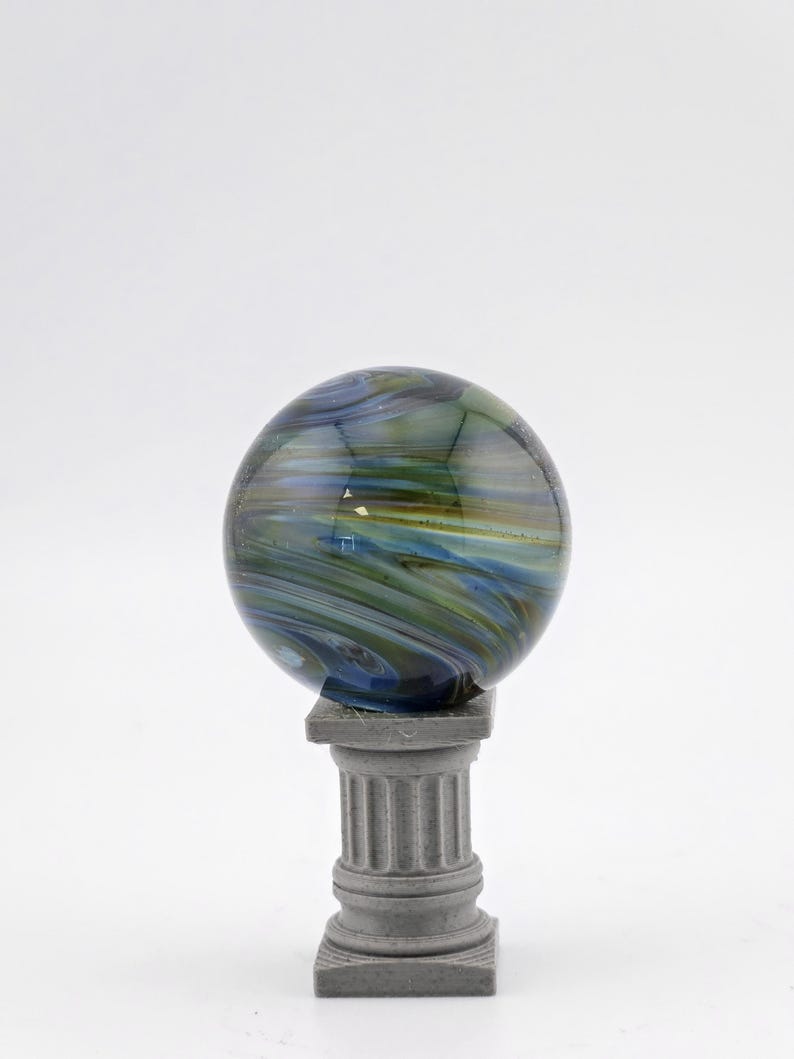 May include: A decorative glass orb with swirling blue, green, and yellow colors, resting on a small, gray, classical-style column. The orb has a polished, reflective surface, and the column has fluted detailing.