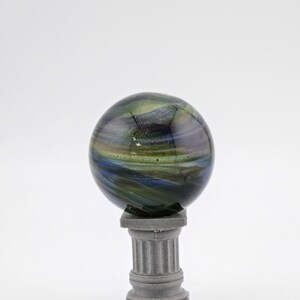 May include: A decorative glass sphere with swirling blue, green, and gold colors, resting on a small, gray, classical-style pedestal. The sphere has a glossy finish and appears to be made of fused glass. The pedestal is a miniature column.