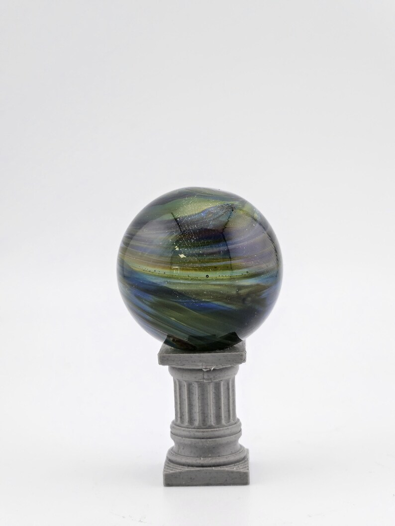May include: A decorative glass sphere with swirling blue, green, and gold colors, resting on a small, gray, classical-style pedestal. The sphere has a glossy finish and appears to be made of fused glass. The pedestal is a miniature column.