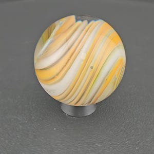 May include: A spherical glass marble featuring a swirling pattern of opaque white, translucent yellow, and light amber bands. The surface is smooth and reflective, showcasing a continuous flow of colors that wrap around the entire orb shape.
