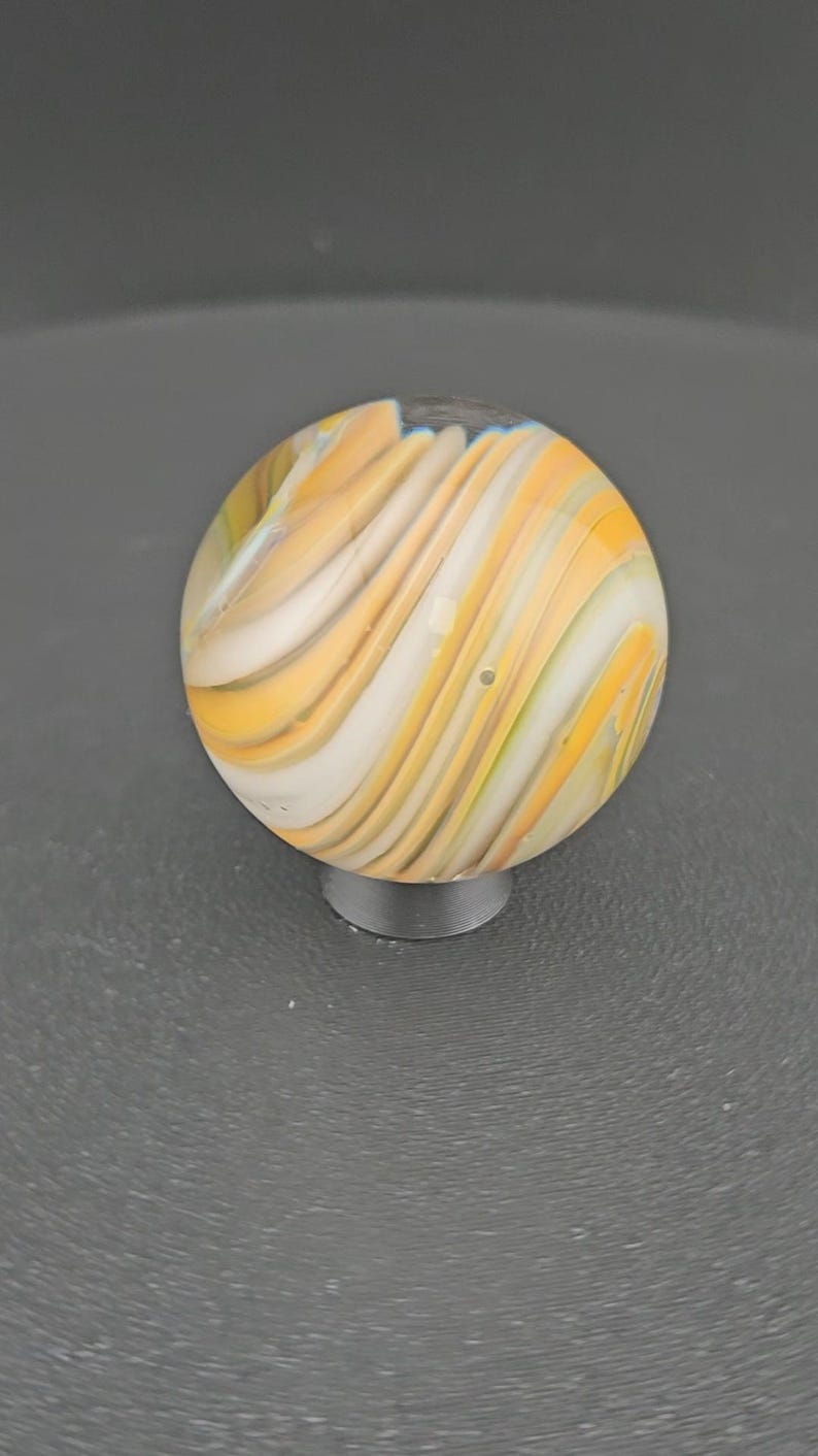 May include: A spherical glass marble featuring a swirling pattern of opaque white, translucent yellow, and light amber bands. The surface is smooth and reflective, showcasing a continuous flow of colors that wrap around the entire orb shape.