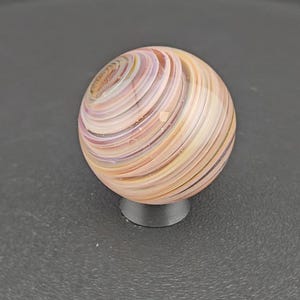 May include: A spherical glass marble featuring swirling bands of soft peach, cream, and lavender colours. The surface is smooth and polished, displaying a continuous spiral pattern that wraps around the entire orb, resting on a small black stand.
