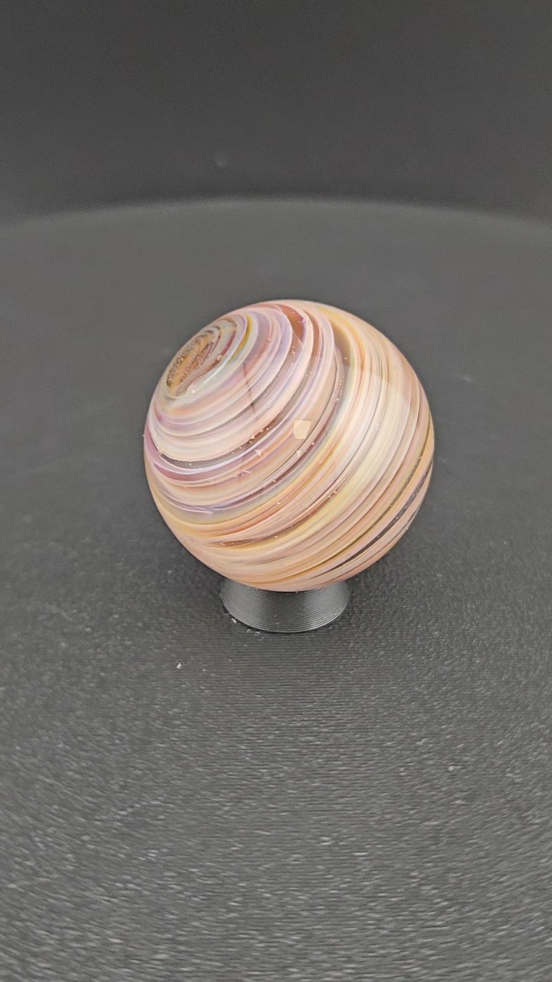 May include: A spherical glass marble featuring swirling bands of soft peach, cream, and lavender colours. The surface is smooth and polished, displaying a continuous spiral pattern that wraps around the entire orb, resting on a small black stand.