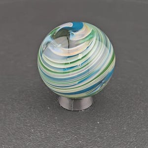 May include: A spherical glass marble featuring swirling patterns of blue, green, white, and translucent clear glass. The marble is resting on a small, circular, dark gray stand that provides a stable base for the rounded object to be displayed.