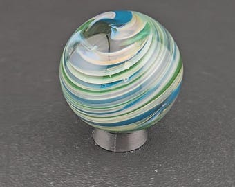May include: A spherical glass marble featuring swirling patterns of blue, green, white, and translucent clear glass. The marble is resting on a small, circular, dark gray stand that provides a stable base for the rounded object to be displayed.