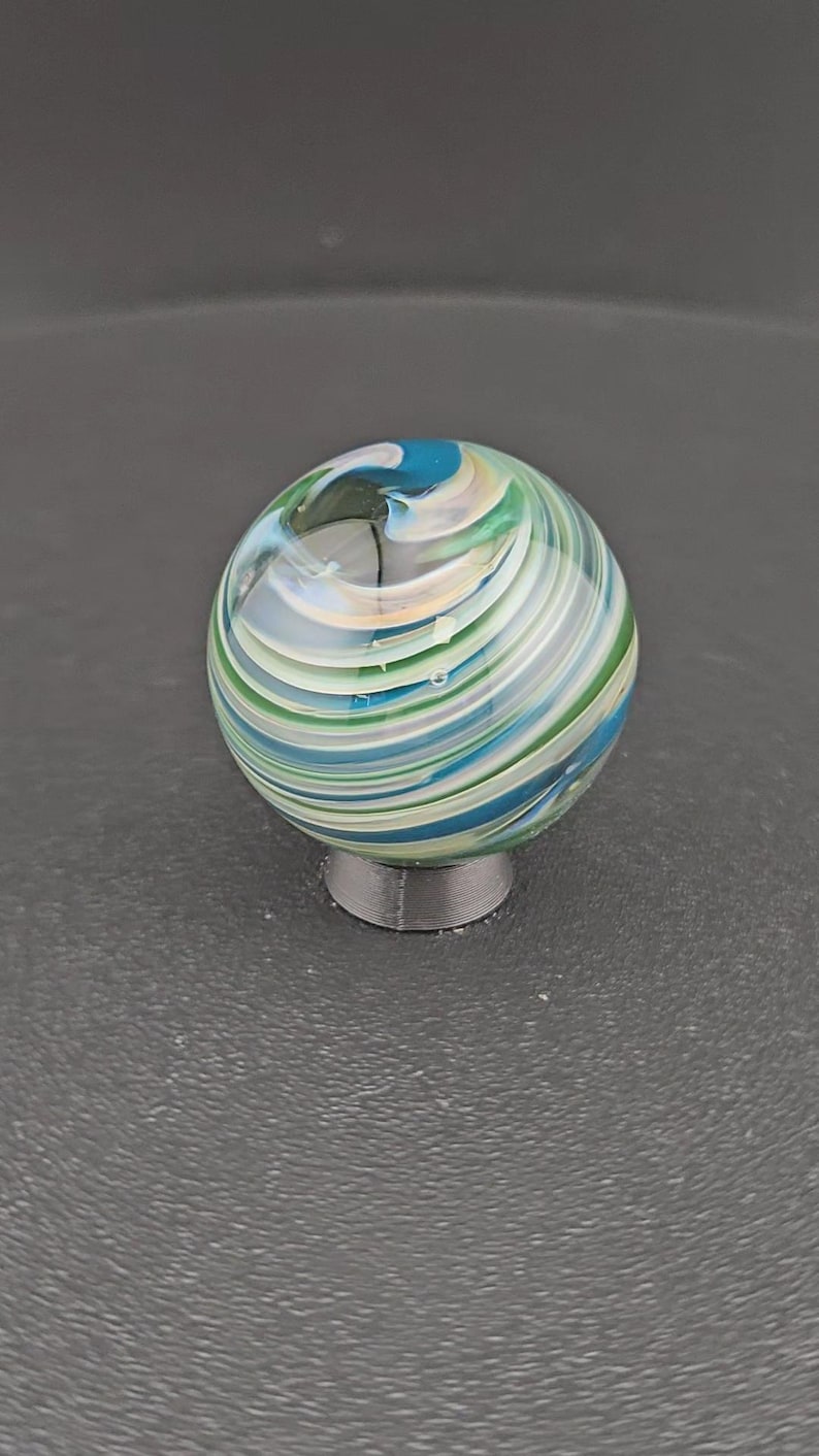 May include: A spherical glass marble featuring swirling patterns of blue, green, white, and translucent clear glass. The marble is resting on a small, circular, dark gray stand that provides a stable base for the rounded object to be displayed.