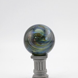 May include: A decorative glass sphere with swirling blue, green, and yellow colors, resting on a small, gray, classical-style pedestal. The sphere has a polished, reflective surface, and the pedestal has a square base and fluted column.
