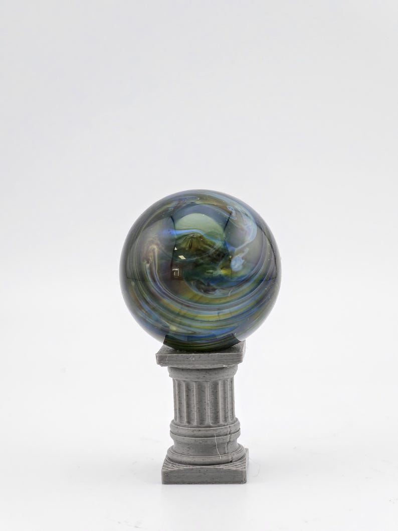 May include: A decorative glass sphere with swirling blue, green, and yellow colors, resting on a small, gray, classical-style pedestal. The sphere has a polished, reflective surface, and the pedestal has a square base and fluted column.
