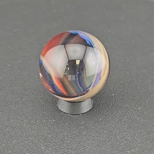 May include: A spherical glass marble featuring swirling patterns of deep blue, vibrant red, and creamy beige colors. The marble is clear and polished, reflecting light on its surface, and is supported by a small, circular, dark gray plastic stand.