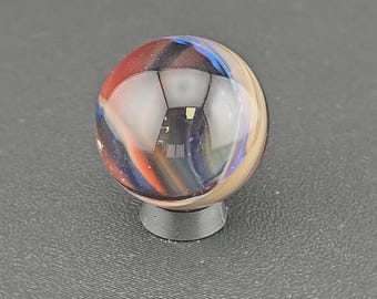 May include: A spherical glass marble featuring swirling patterns of deep blue, vibrant red, and creamy beige colors. The marble is clear and polished, reflecting light on its surface, and is supported by a small, circular, dark gray plastic stand.