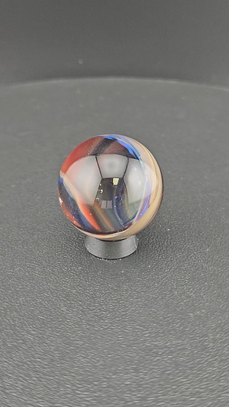 May include: A spherical glass marble featuring swirling patterns of deep blue, vibrant red, and creamy beige colors. The marble is clear and polished, reflecting light on its surface, and is supported by a small, circular, dark gray plastic stand.