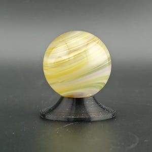 May include: A decorative glass sphere with yellow, white, and green swirls, resting on a black, spiral-shaped base. The sphere has a polished, glossy finish. The base is made of a dark material.