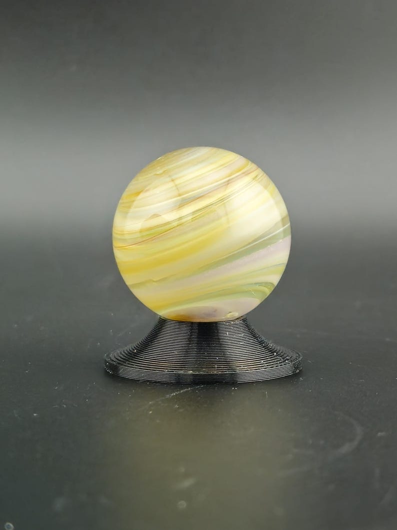 May include: A decorative glass sphere with yellow, white, and green swirls, resting on a black, spiral-shaped base. The sphere has a polished, glossy finish. The base is made of a dark material.