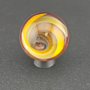 May include: A spherical glass marble featuring a swirling internal pattern of bright yellow and translucent white ribbons encased within a clear outer layer. The marble is positioned on a small, dark, circular base, showcasing a smooth, polished surface.