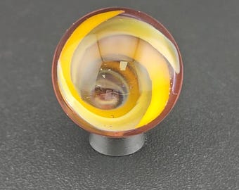 May include: A spherical glass marble featuring a swirling internal pattern of bright yellow and translucent white ribbons encased within a clear outer layer. The marble is positioned on a small, dark, circular base, showcasing a smooth, polished surface.